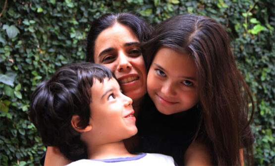 Mulher abraça duas crianças, um menino pequeno de cabelos castanhos curtos e uma menina de cabelos longos e lisos. Os três sorriem próximos uns dos outros, em frente a um muro coberto por folhagem verde.