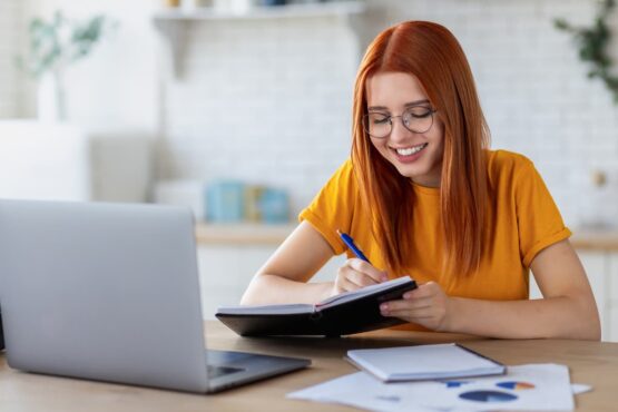 Imagem de uma mulher ruiva com blusa laranja estudando na frente de um computador