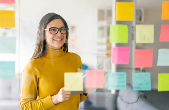 Imagem de uma mulher sorridente em frente a um quadro para organizar tarefas cheio de post-it.