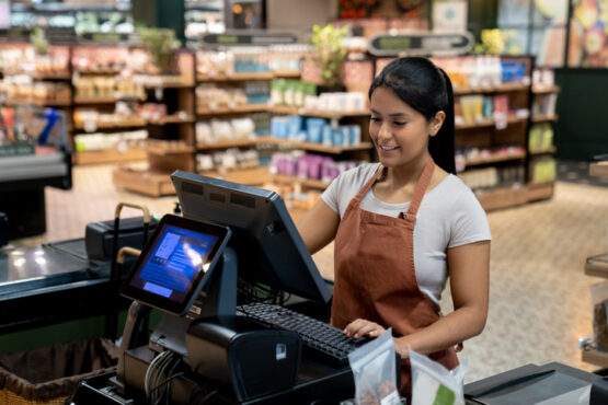 Funcionária de supermercado sorrindo enquanto opera o caixa em frente a um computador e teclado, com prateleiras de produtos ao fundo.