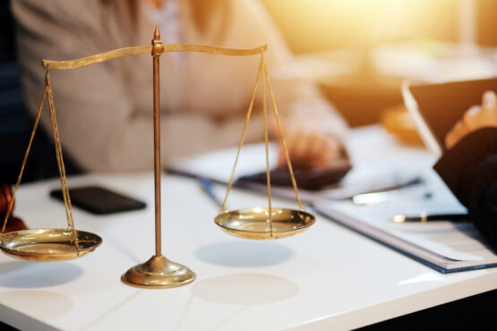 Balança dourada da justiça sobre mesa de escritório com documentos, ao fundo pessoas em reunião.